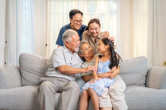 Portrait of Happy Asian three generation family spending time together at home. Grandparents hugging grandchild girl on sofa in living room. Big family enjoy relaxing and weekend activities lifestyle.