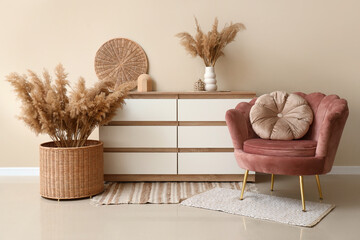 Armchair, chest of drawers and wicker basket with pampas grass near white wall in room