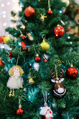 Fototapeta premium Toy angel hangs on a branch of a Christmas tree among colored balls, bells and luminous garlands