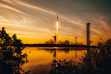 Rocket launch at sunset with fiery trail over water, reflected in golden hues. SpaceX Falcon rocket...