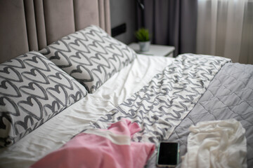 Bedroom Bedding Clothes Mess: Unmade bed, discarded clothes, smartphone; modern bedroom interior, daytime, likely after sleep.