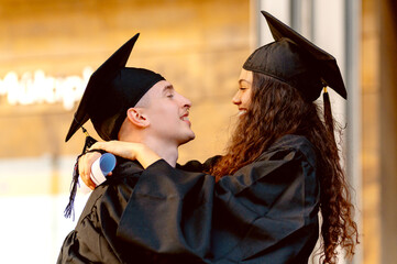 Two happy graduates wearing mortarboards and gowns are embracing each other at their graduation...