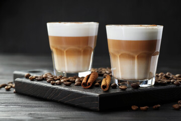 Tasty latte macchiato in glasses, coffee beans and cinnamon on black wooden table, closeup