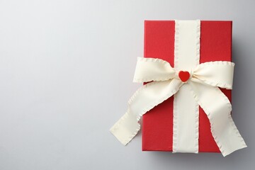 Gift box with beige bow and decorative heart on light grey background, top view. Space for text