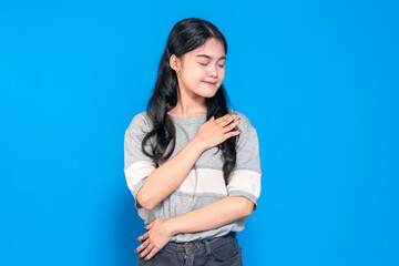 Young Asian woman with long black hair gently smiling and hands on her chest, expressing gratitude and serenity. She wears a casual outfit against a blue background.