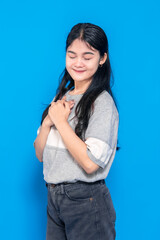 Obraz premium Young Asian woman with long black hair gently smiling and hands on her chest, expressing gratitude and serenity. She wears a casual outfit against a blue background.