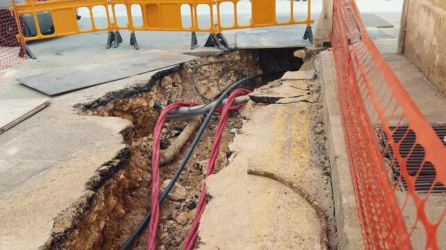 A deep trench with exposed underground cables and pipes on a damaged asphalt road. Construction barriers and safety nets surround the site. Infrastructure repair and urban development in progress.