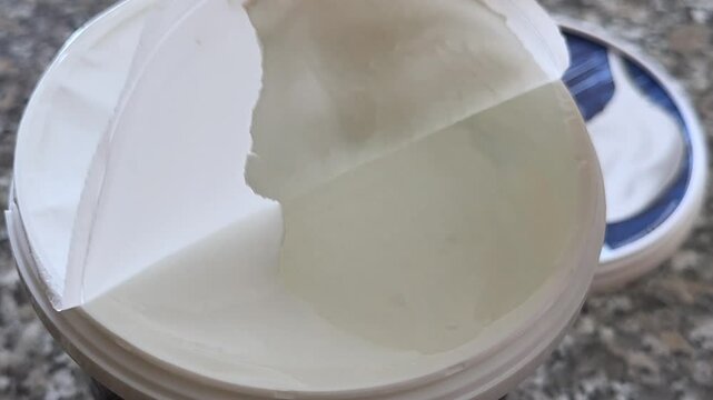 Unpacking the product : a hand removes the protective foil lid from a jar of natural yogurt, revealing its fresh, creamy content. The shot focuses on the yogurt&rsquo;s texture and presentation.