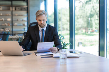 Mature businessman with beard working with documents, contracts and bills sitting at table using laptop at work in modern bright office with large windows
