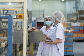 Two female workers wearing protective clothing and face masks conduct a quality control inspection...