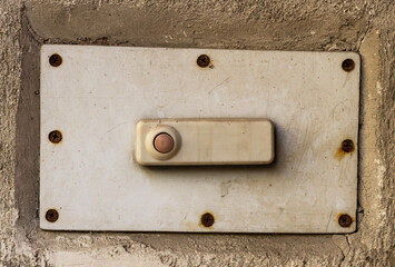 An old-fashioned doorbell button, mounted on a wall. The button itself is an elongated oval shape, slightly raised, with a small circular button in the center. 