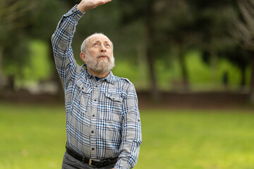 Senior man stretching arm and protecting eyes from sunlight in park