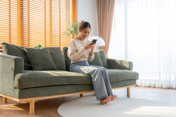 Happy young asian woman relax on comfortable couch at home texting messaging on smartphone, smiling...