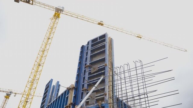 Construction site with tall concrete formwork structure. Metal reinforcements extend from framework and cranes stand on solid foundation