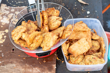 Crispy fried tempeh with a seasoned coating in a strainer above cardboard and a blue surface. Some pieces are in a clear plastic container