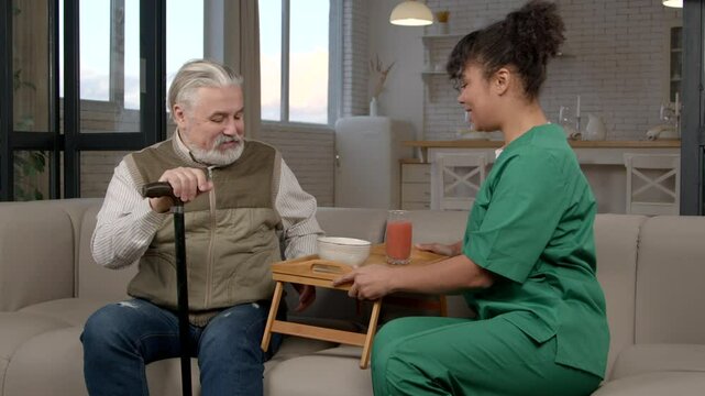 Lovely black female carer in uniform helping elderly grey haired man on retirement with walking stick with activities of daily living, bringing healthy breakfast in nursing home.