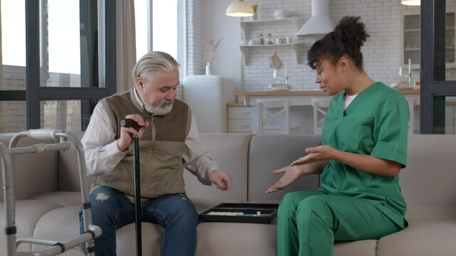 Positive handsome senior grey haired man holding walking cane , playing backgammon game with pretty black female caregiver on sofa, rolling dices and moving checkers in care home.