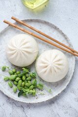 Freshly cooked chinese baozi with edamame beans on a grey plate, vertical shot on a white stone background, elevated view