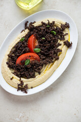 Hummus served with fried ground beef and tomato wedges, vertical shot on a light-beige stone background, high angle view
