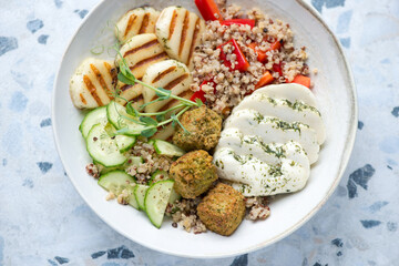 Halloumi cheese with quinoa, falafel and fresh vegetables served in a beige plate, horizontal shot on a white and blue granite surface, middle close-up