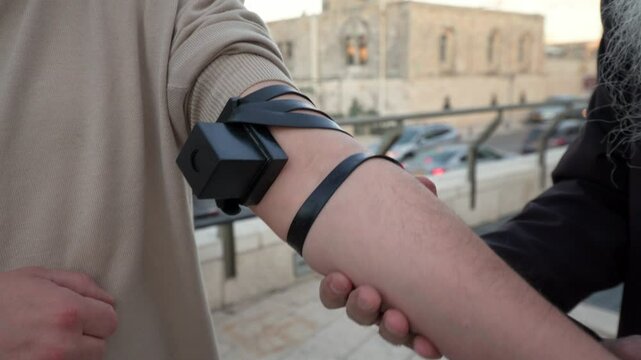 Rabbi's hands wrap tefillin on a Jewish man's arm against the backdrop of Jerusalem. A sacred ritual symbolizing faith, devotion, and Jewish tradition in the holy city. Spiritual and cultural heritage