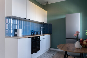 design of modern small kitchen. built-in kitchen with white cabinets, blue tile backsplash, wooden countertop. white kitchen unit. steel refrigerator, oven, flowers on vase, white faucet