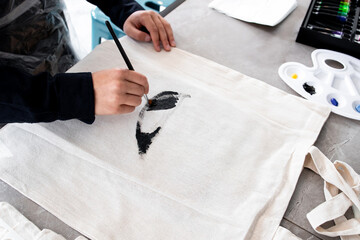 Person painting design on canvas bag with black brush strokes in art studio
