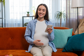 Smiling pregnant woman using smartphone text messaging social media chatting sitting on sofa in...
