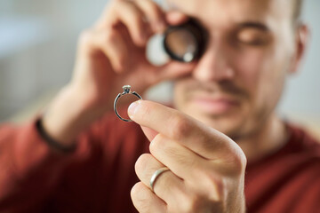 Closeup portrait of young man jeweler looking through magnifying glass examining gold ring...