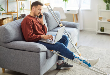 Young man with broken leg working on laptop at home talking on mobile phone sitting on sofa with...