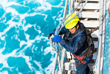 A deckhand prepares the pilot ladder as the ship readies for port entry. Secured in a safety harness, he carefully ensures the setup is ready, focusing on the task in a high-risk environment. 