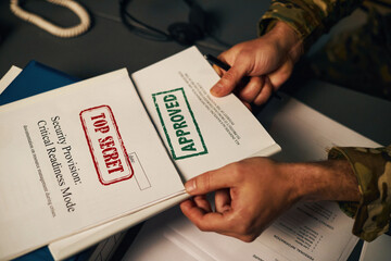 Person's hands holding documents labeled top secret and approved at desk with scattered papers,...