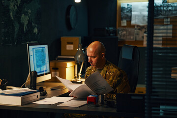 Man in military uniform analyzing classified documents at his desk, surrounded by computers and...