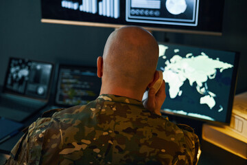 Person in camouflage uniform analyzing data on multiple screens in a control room. World maps on display indicating geopolitical hotspots and security threats