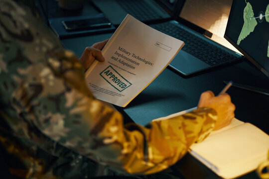 Soldier reviewing military technologies document on desk with computer screens displaying maps and data. Engaging in military documentation in office setting