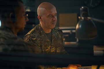 Two military personnel engaging in strategic planning at dimly lit meeting room. Camouflaged uniforms and serious expressions emphasize their focus and dedication