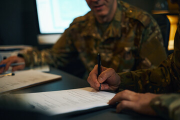 Military personnel signing official documents during work in a dimly lit office environment,...