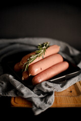 Raw sausages with rosemary and pepper in a frying pan on a wooden board on a black background