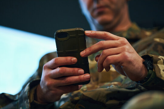Military personnel in camouflage uniform closely handling smartphone in hand, highlighting modern communication and technology in use. Partial face visible in background