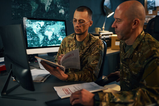 Military personnel in camouflage uniforms reviewing classified documents in front of computer monitors in command center. Engaged in serious discussion and analysis in secure environment