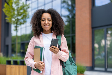 A smiling college student stands outside of a building holding a phone and notebooks. She has a backpack slung over her shoulder.