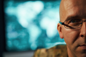 Portrait of man looking intently at camera, wearing military uniform with glasses and short hair. Background displays blurred digital screen, creating focused atmosphere