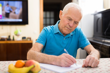 Serious older man sitting at table writing letter or filling some paper form