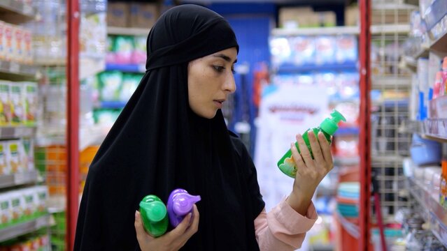 Young muslim woman wearing a black hijab is comparing two liquid soap bottles in a supermarket, carefully examining the green one while holding a purple one