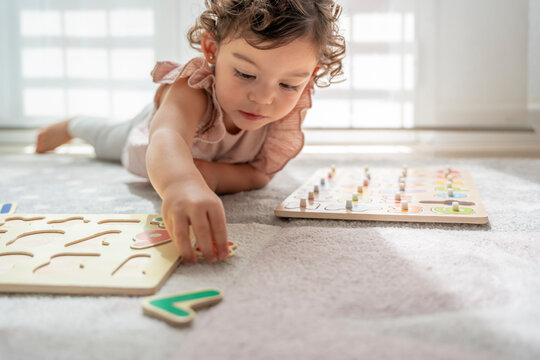 Horizontal photograph of a little girl lying down playing with wooden letter and number puzzles. Fun and learning in childhood. Development of cognitive and motor skills. Montessori education.