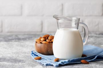 Glass milk jug resting on blue napkin beside wooden bowl filled with almonds, gray kitchen table featuring white brick wall backdrop, highlighting nutritious beverage