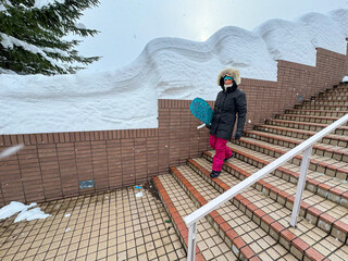 Cheerful Caucasian woman dressed in a warm black coat and pink snow pants carries a snowboard while...
