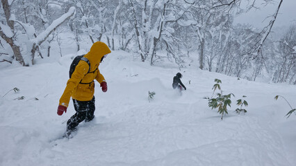 Two unrecognizable extreme snowboarders carve through Hokkaido’s deep powder. One wears a yellow...