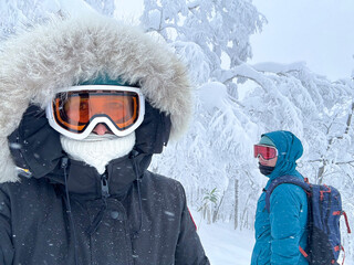 SELFIE: Snowboarders dressed in heavy winter gear with goggles and insulated clothing, explore a snow-covered forest. The icy trees and falling snow create a strikingly frozen, adventurous atmosphere.