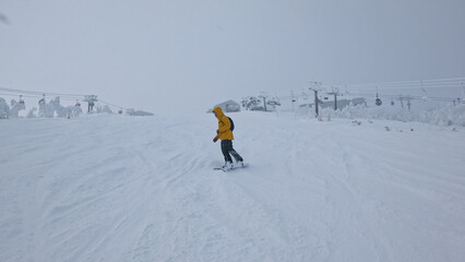 Active young man in yellow ski coat snowboards down a ski resort slope on a cloudy winter day in...